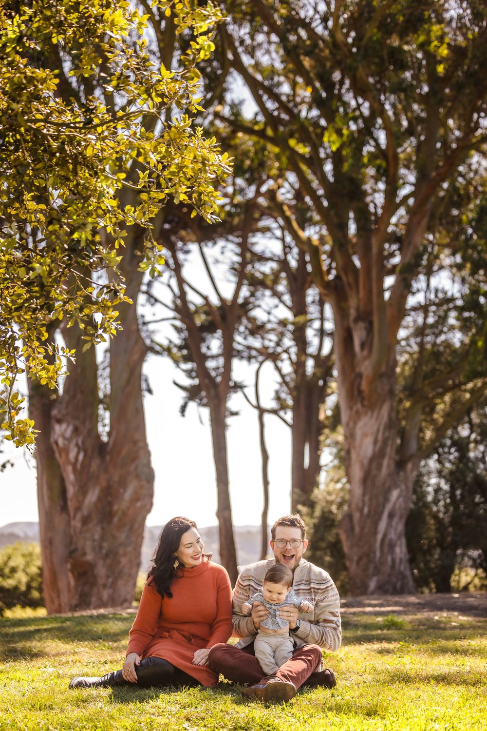 Family sitting on grass under large trees in a sunny park, parents smiling at baby in warm autumn colors.