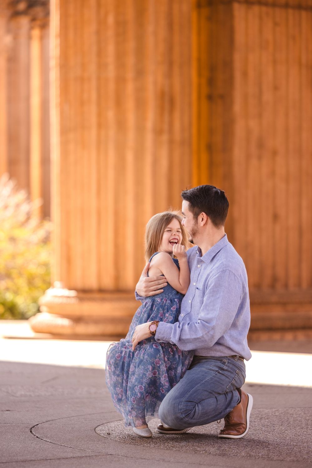 A father kneels to embrace his laughing young daughter in a blue floral dress near golden columns.