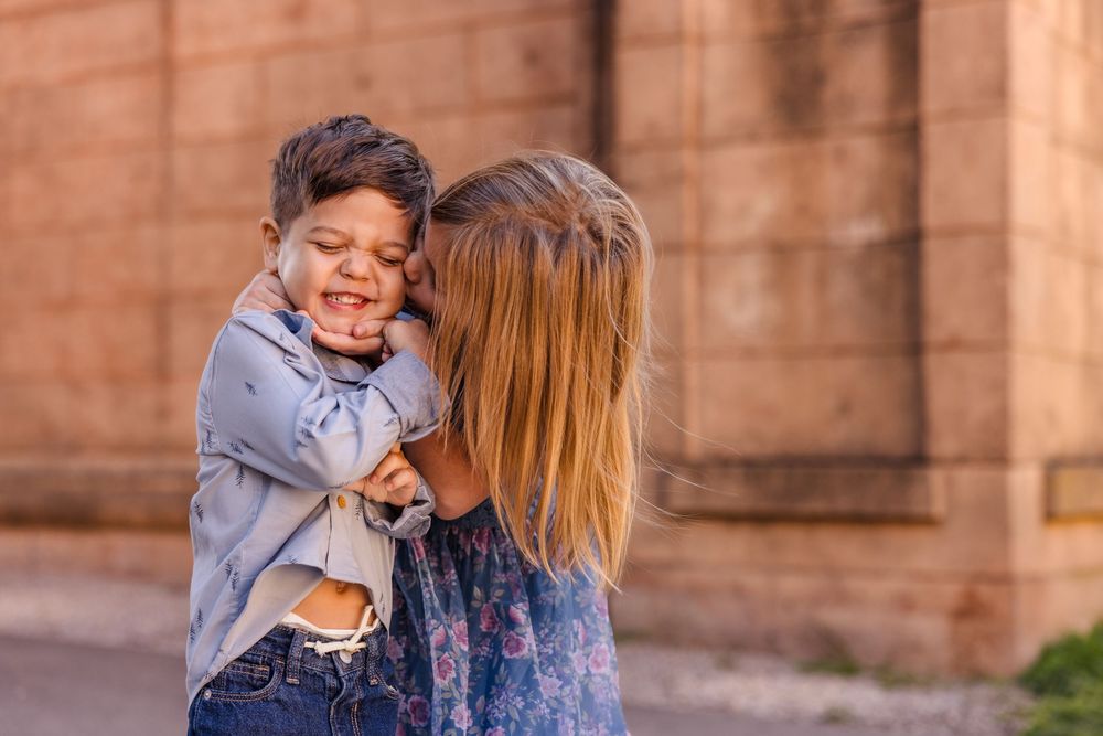 A joyful young boy laughs while being hugged by a woman with long blonde hair outdoors near a brick wall.