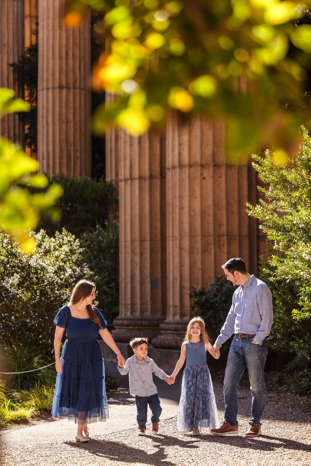A happy family of four holds hands while walking together outdoors near large stone columns in warm sunlight.