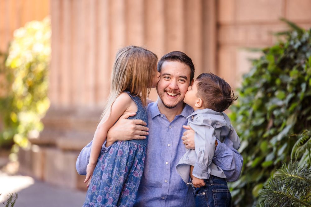 A smiling man holds two young children, a girl kissing his cheek and a baby, outdoors near greenery.
