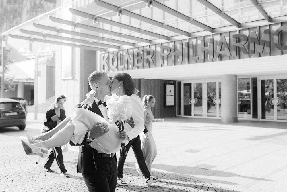 Groom carrying bride outside Kölner Philharmonie concert hall in a romantic black and white wedding photo.