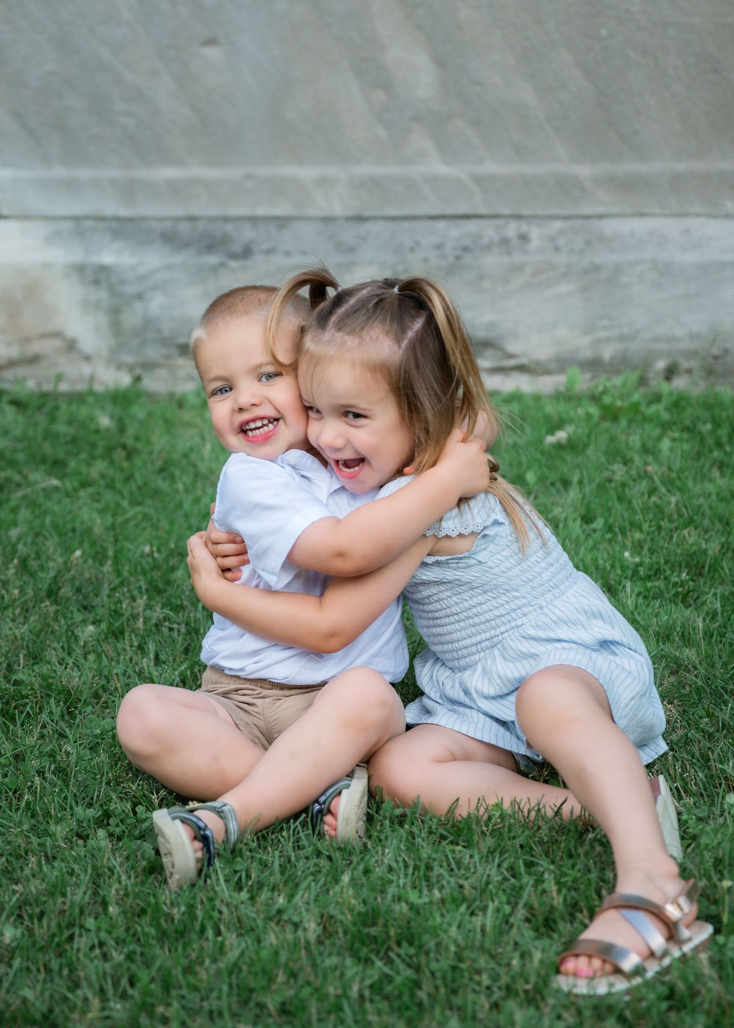 Two young children hugging and laughing together while sitting on green grass outdoors.