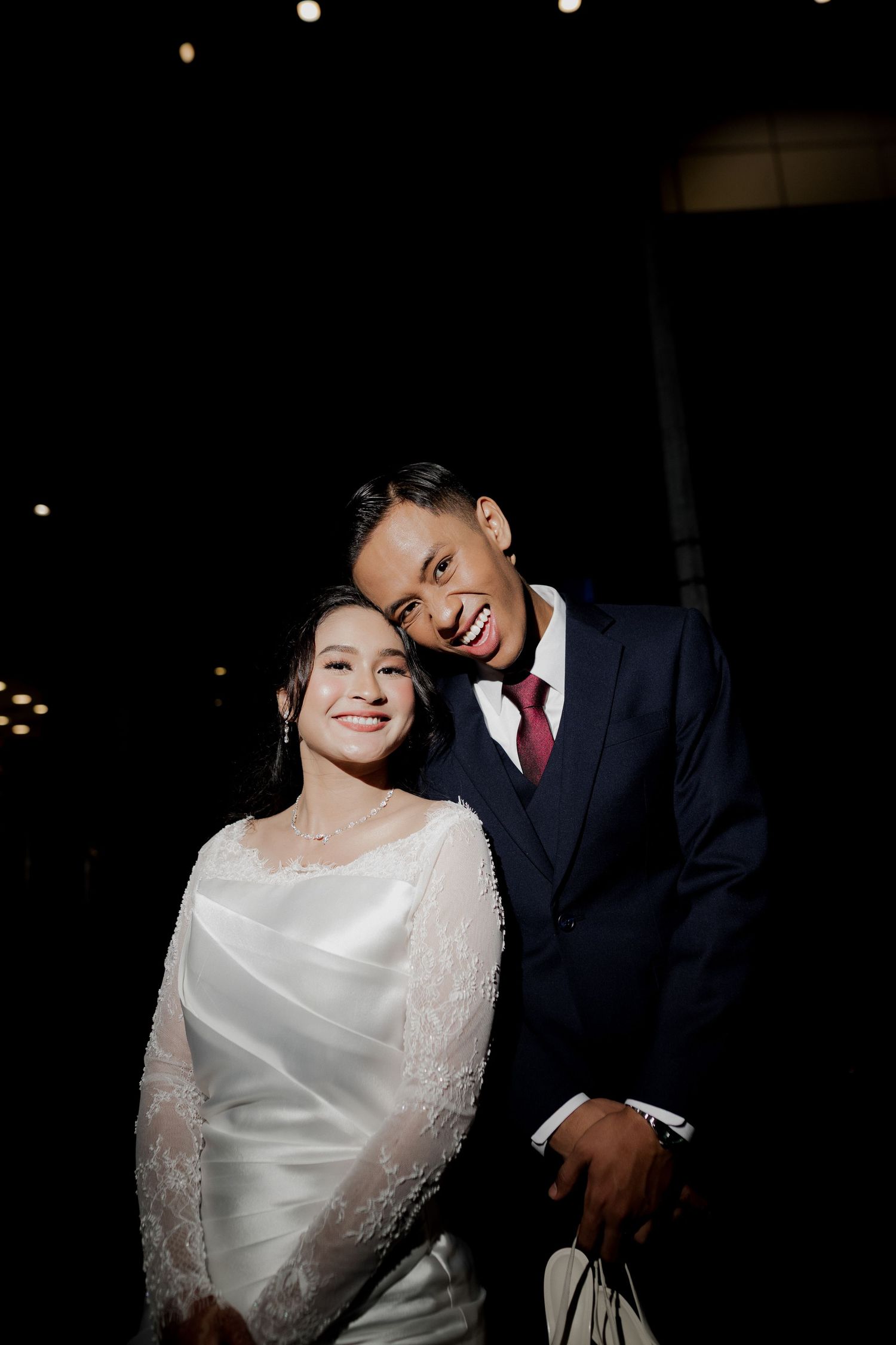 A smiling bride in white lace gown and groom in navy suit with red tie pose together at a nighttime wedding.