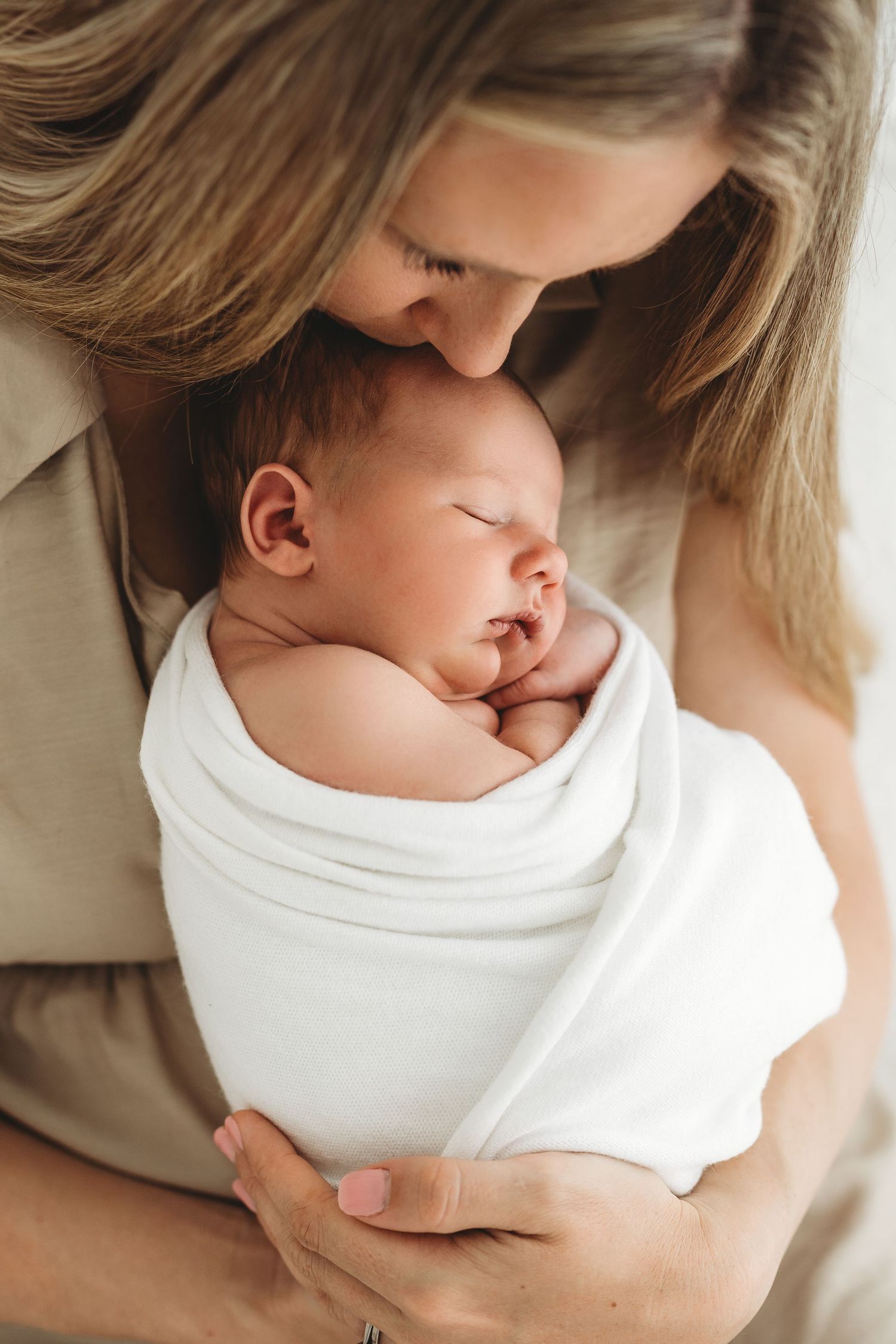 Blonde mother tenderly kissing sleeping newborn baby wrapped in white swaddle blanket close-up portrait.