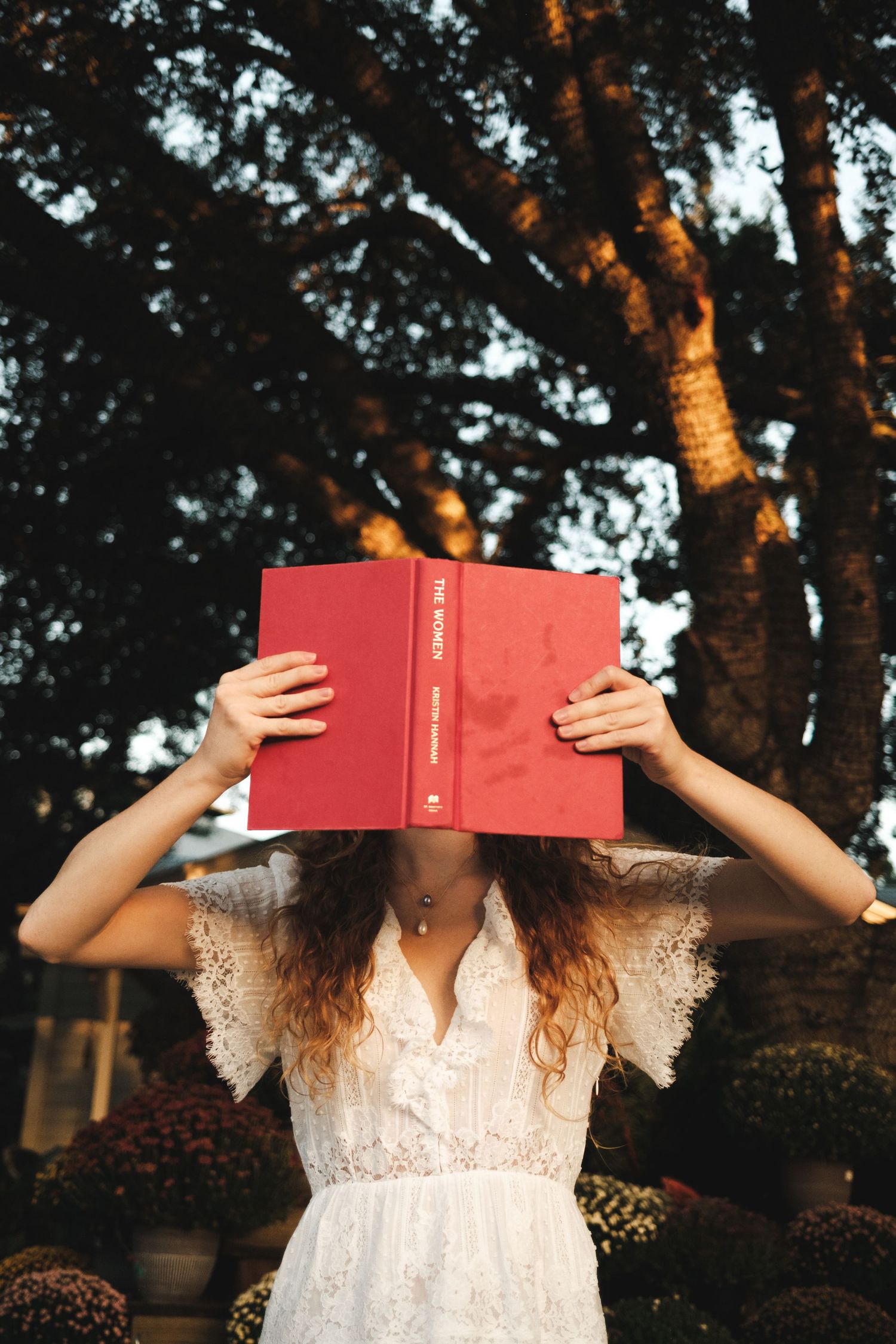 Woman in white lace dress holds red hardcover book over her face outdoors near trees and flowers.