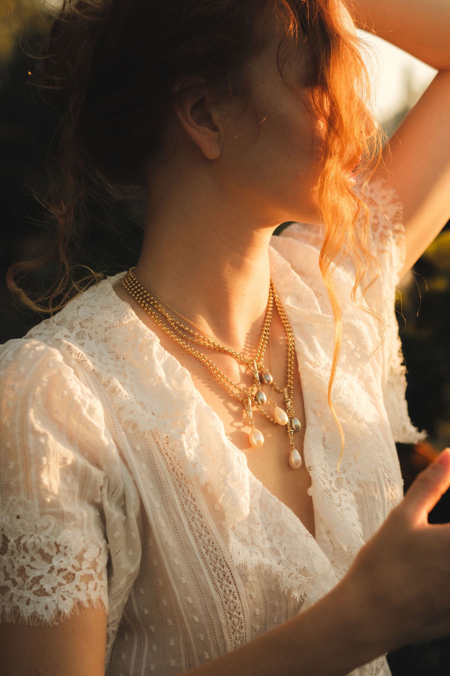 Woman in white lace dress wearing layered gold necklaces with pearl pendants, bathed in warm golden sunlight.