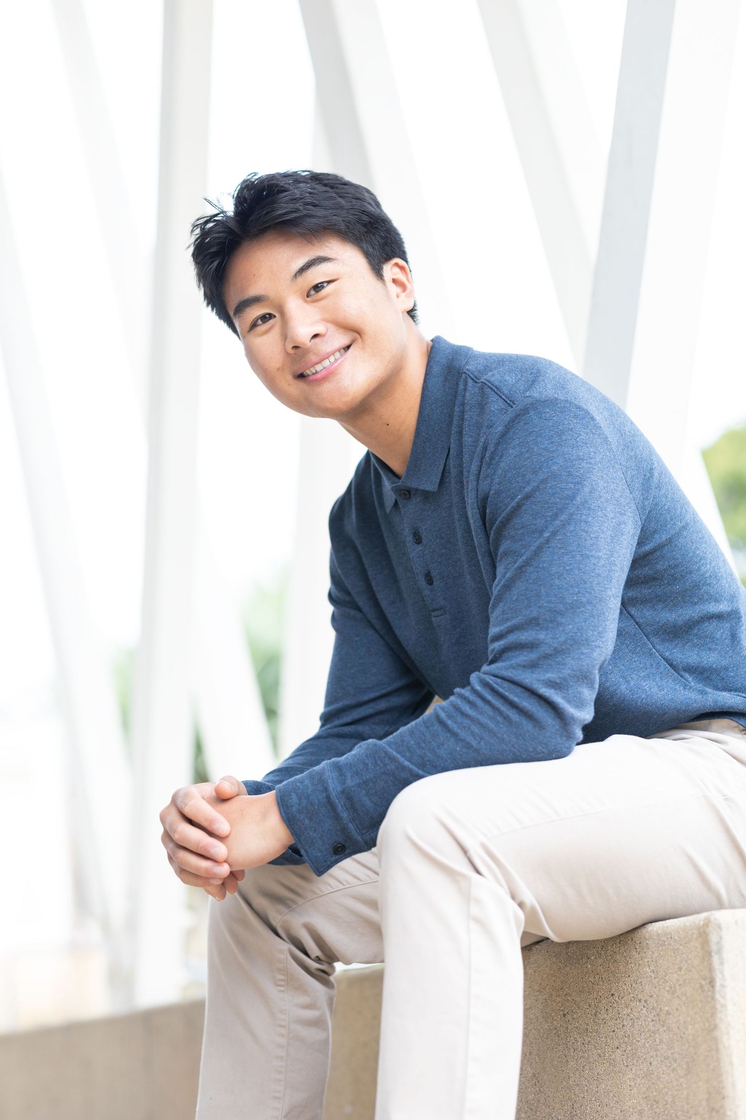Smiling young Asian man in blue sweater sitting relaxed with clasped hands against white architectural background.