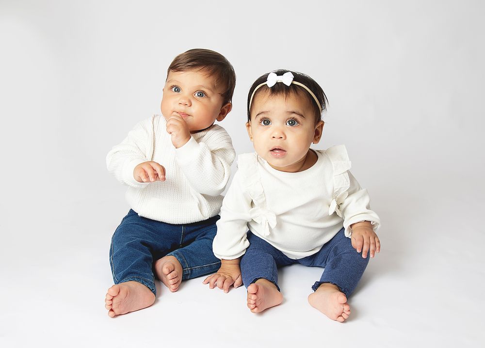 Two cute babies in white tops and jeans sitting together on white background, one with a bow headband. During a family photoshoot in Bexley