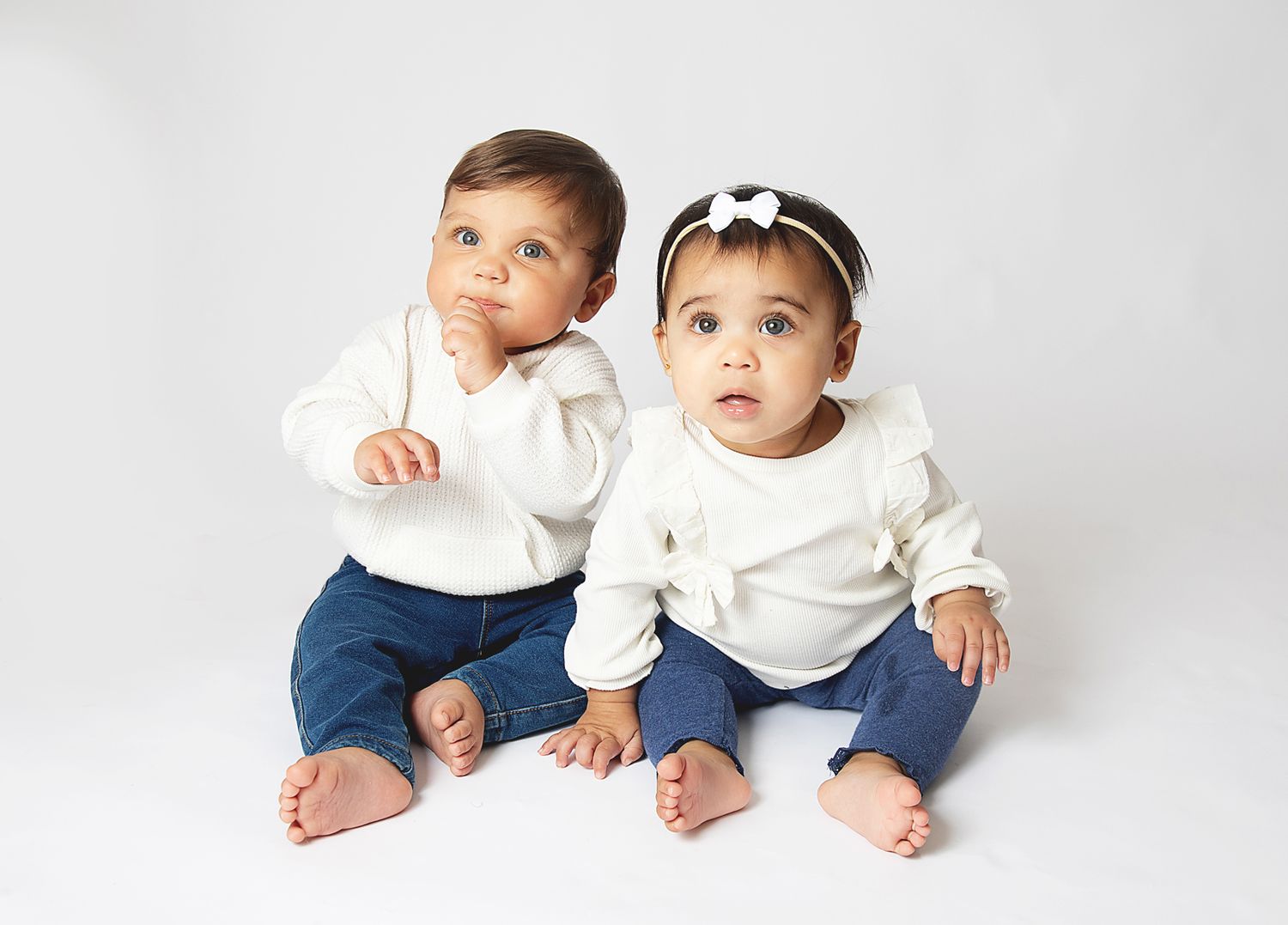 Two cute babies in white tops and jeans sitting together on white background, one with a bow headband. During a family photoshoot in Bexley