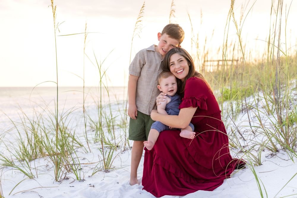 A mother in a red dress sits on white sand with her baby and older son, surrounded by sea oats at sunset.