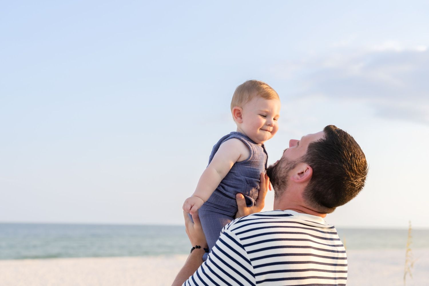 Father lifting smiling baby at beach, ocean in background.