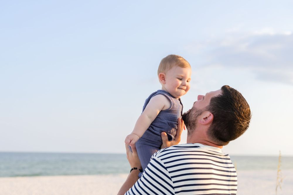 Father lifting smiling baby at beach, ocean in background.