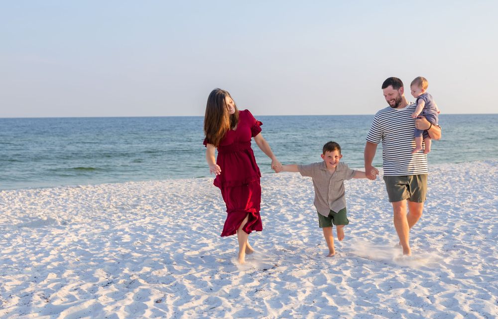 A happy family of four walks on a white sandy beach, with a woman in a red dress, a man holding a toddler.