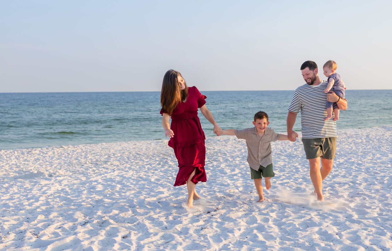 A happy family of four walks on a white sandy beach, with a woman in a red dress, a man holding a toddler.