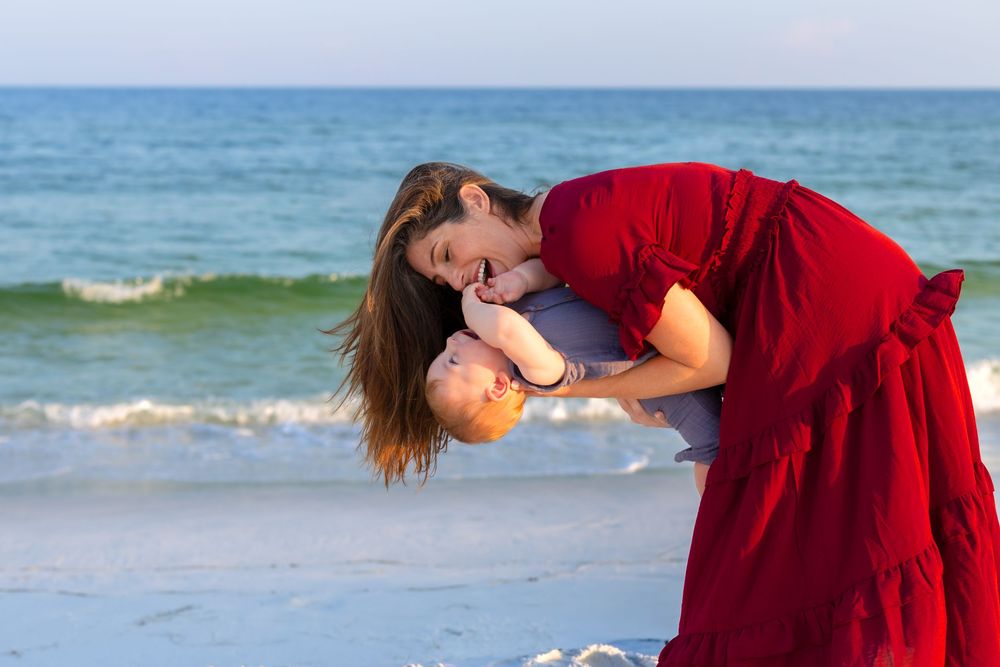 A woman in a red dress joyfully holds a baby on a sunny beach with turquoise ocean waves in the background in Navarre Beach, Florida.