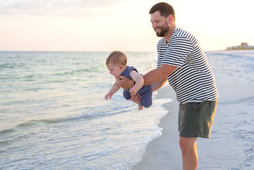Father holding baby over ocean waves on sandy beach at sunset, wearing striped shirt and shorts in Navarre Beach, Florida.