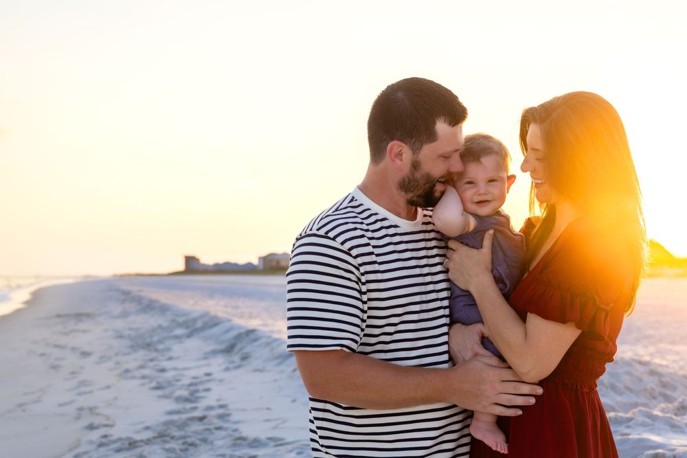 A young family shares a tender moment on a sunny beach, with parents holding their toddler at golden hour.