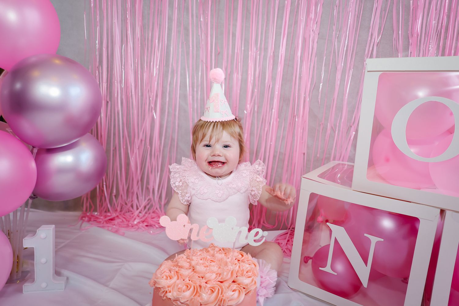 Baby girl in pink birthday hat smiles behind floral cake, surrounded by pink balloons and decorative letter blocks.