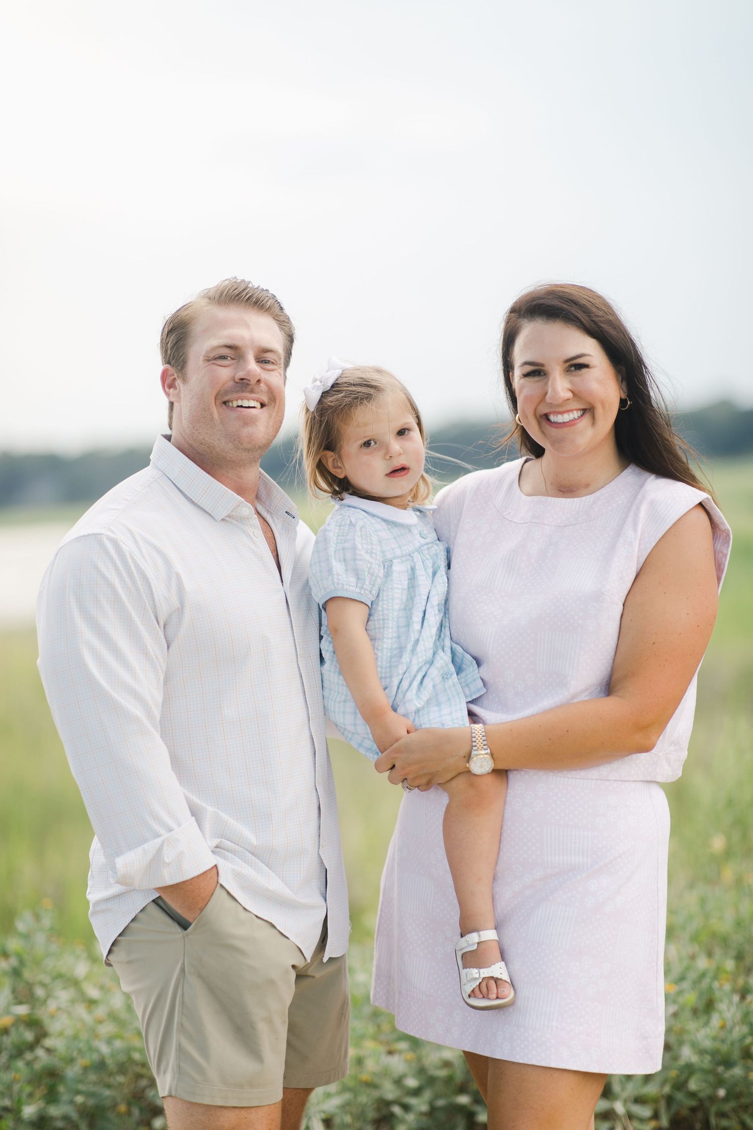 Located in downtown Beaufort, a smiling family of three poses outdoors, with parents in white attire holding a young girl in a blue dress.