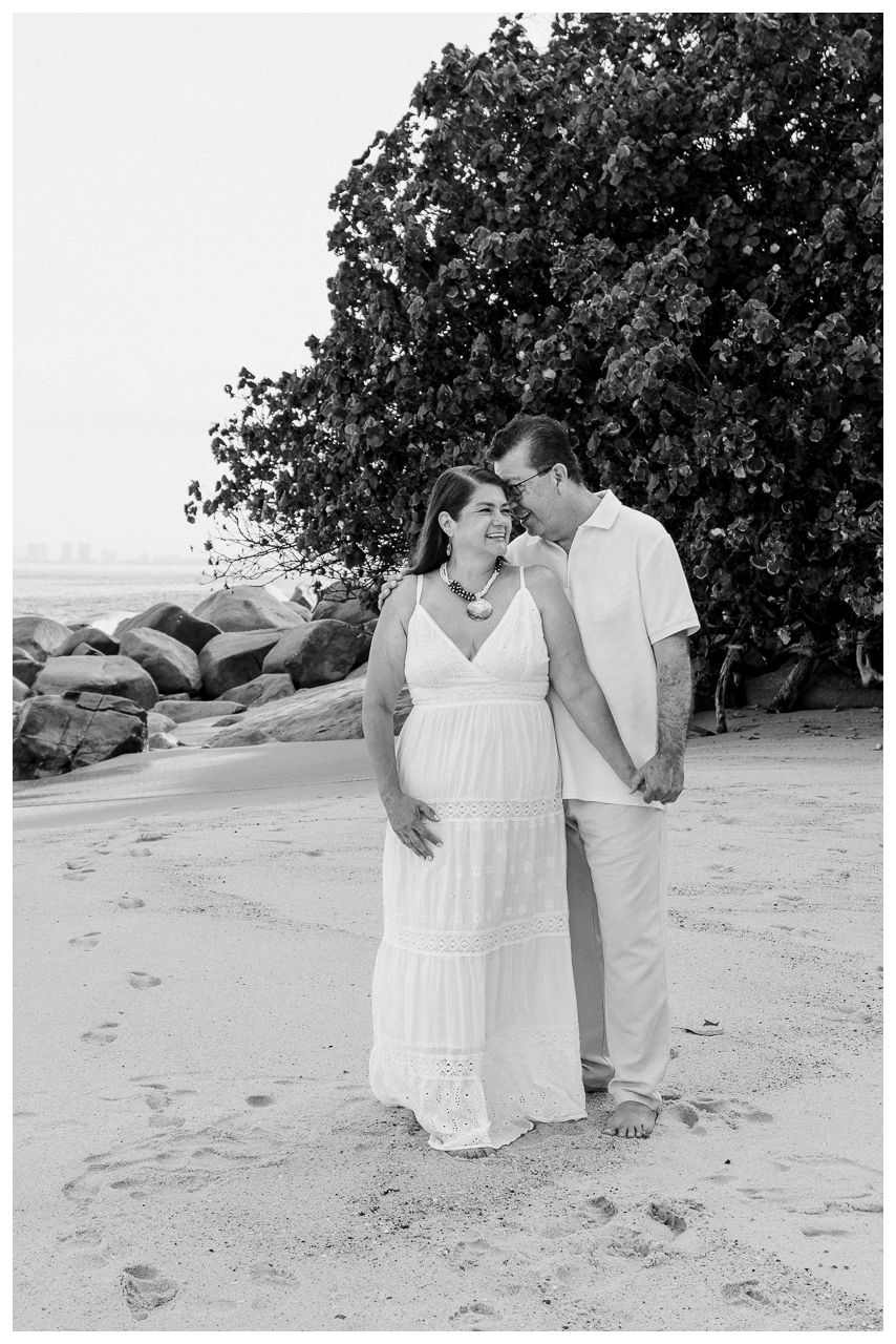 Puerto Vallarta Family Photographer captures Family Portraits of an expecting couple on a sandy beach surrounded by coastal rocks and trees.