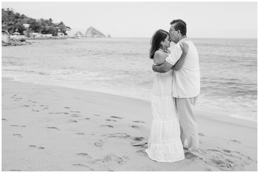 Puerto Vallarta Family Photographer captures Sunset Family Photos of a couple embracing on a sandy beach with ocean waves and rocky cliffs.