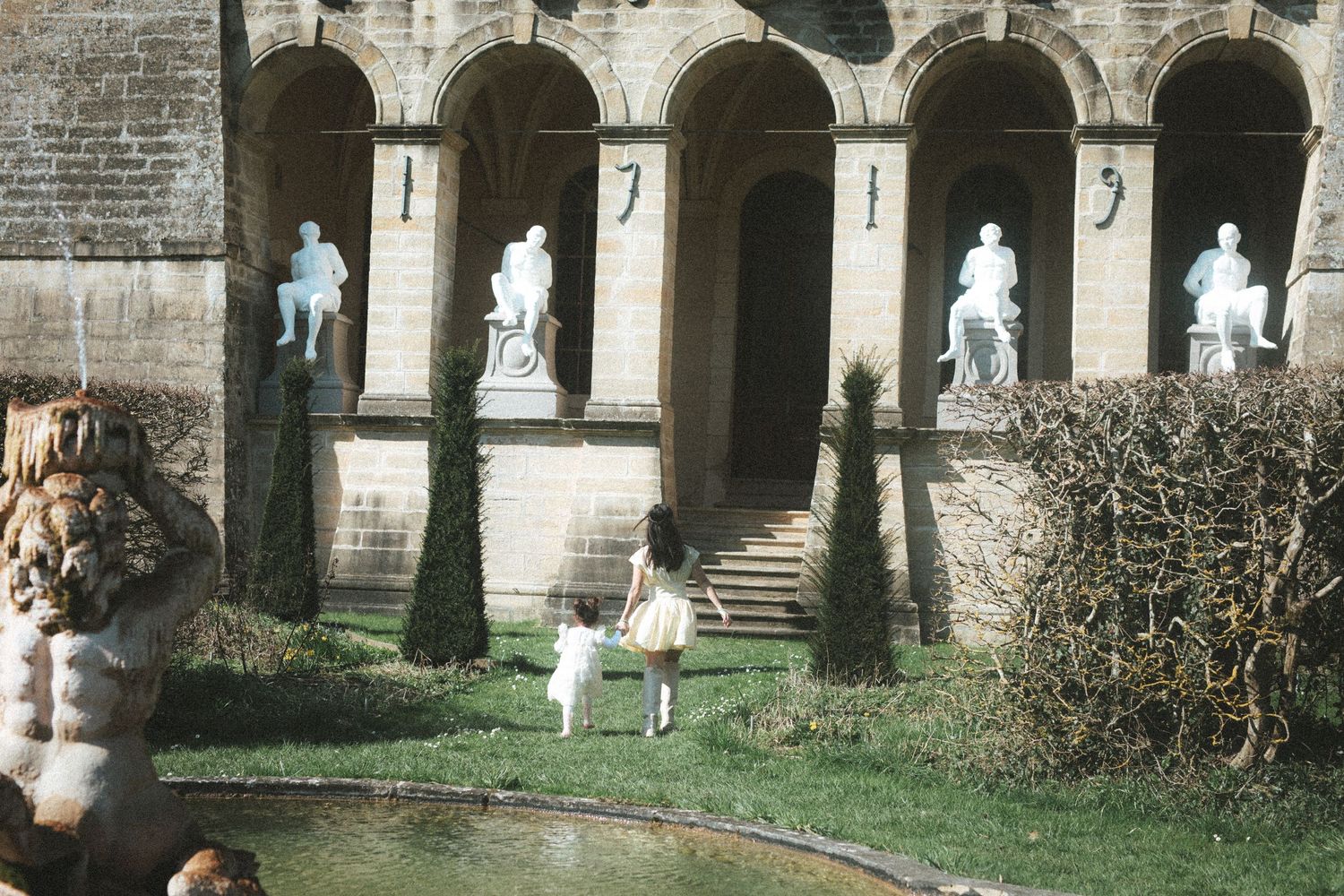 Two children walk toward a grand stone building adorned with classical statues in a formal garden setting.