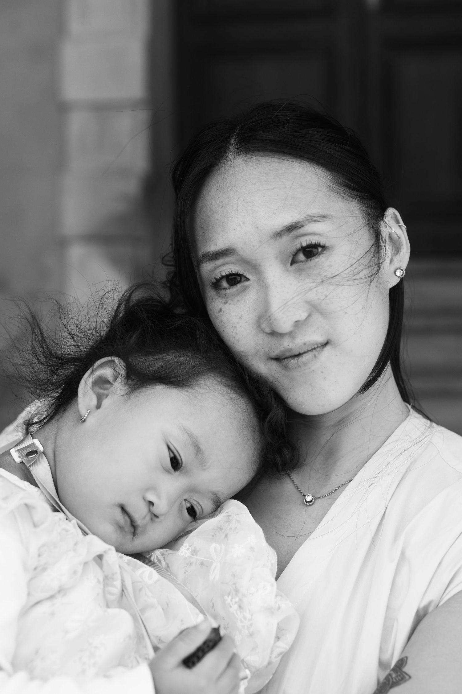 Black and white portrait of an Asian mother tenderly holding her sleeping baby, smiling softly at the camera.