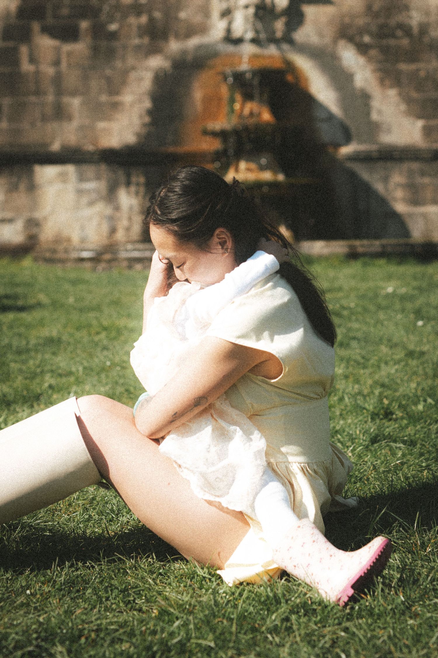 Young woman in white dress and pink boots sitting on grass near a stone fountain in sunlight.