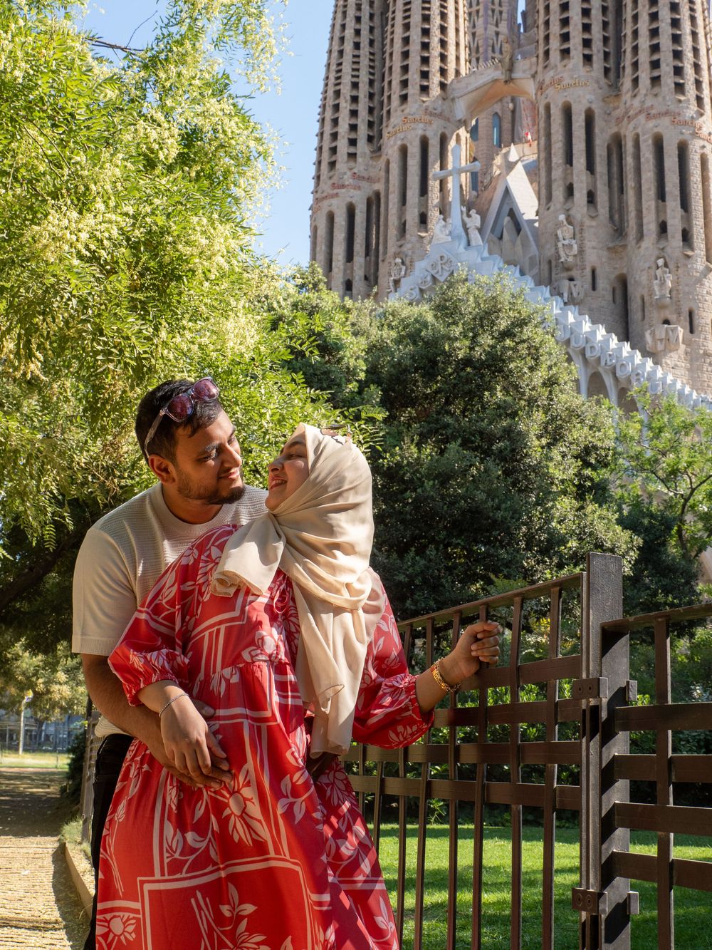 Couple embracing in front of Sagrada Familia basilica in Barcelona, woman in red floral dress and hijab.
