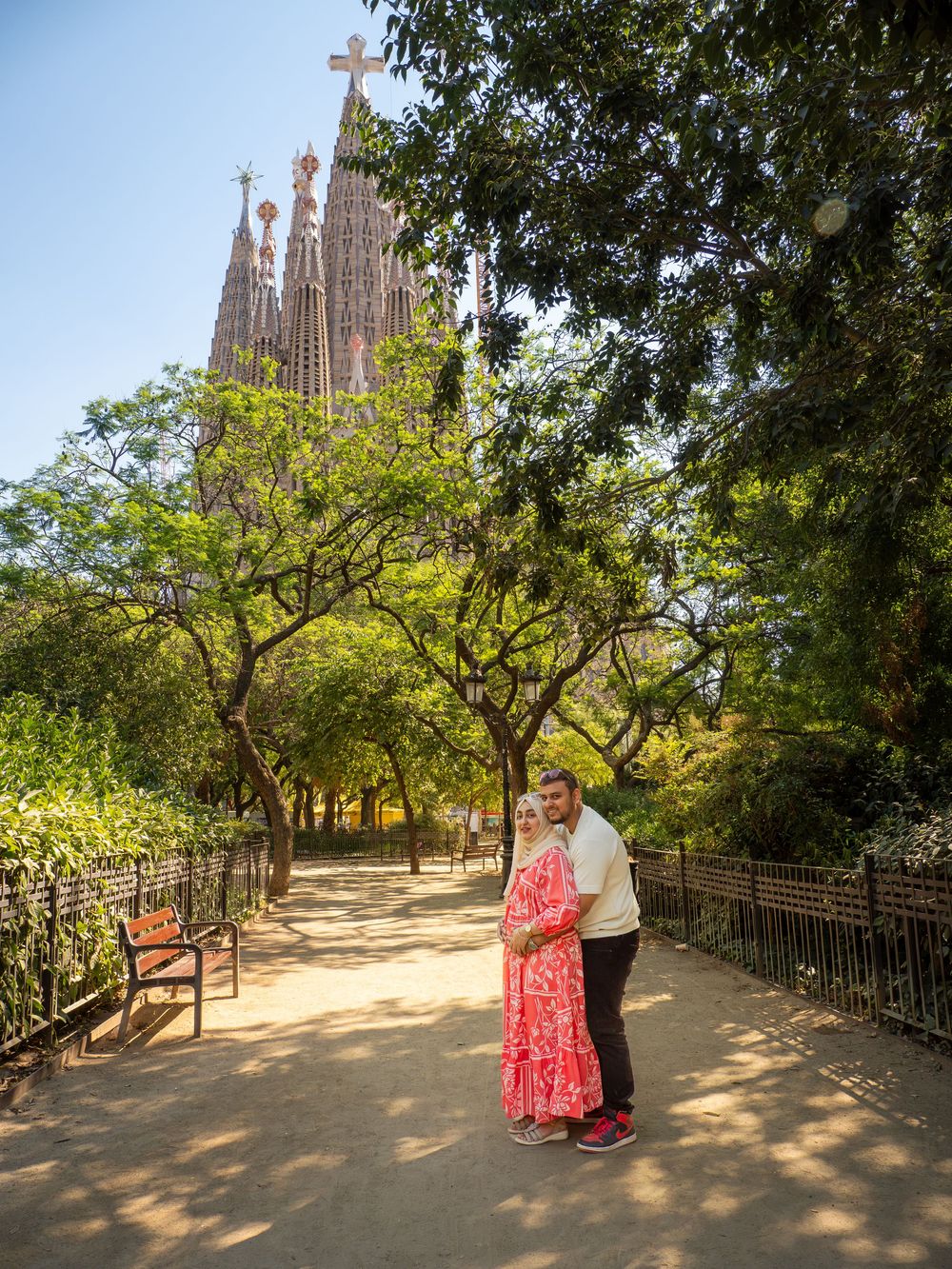 Couple embracing in a park pathway with Sagrada Familia basilica towers visible through lush green trees in Barcelona.