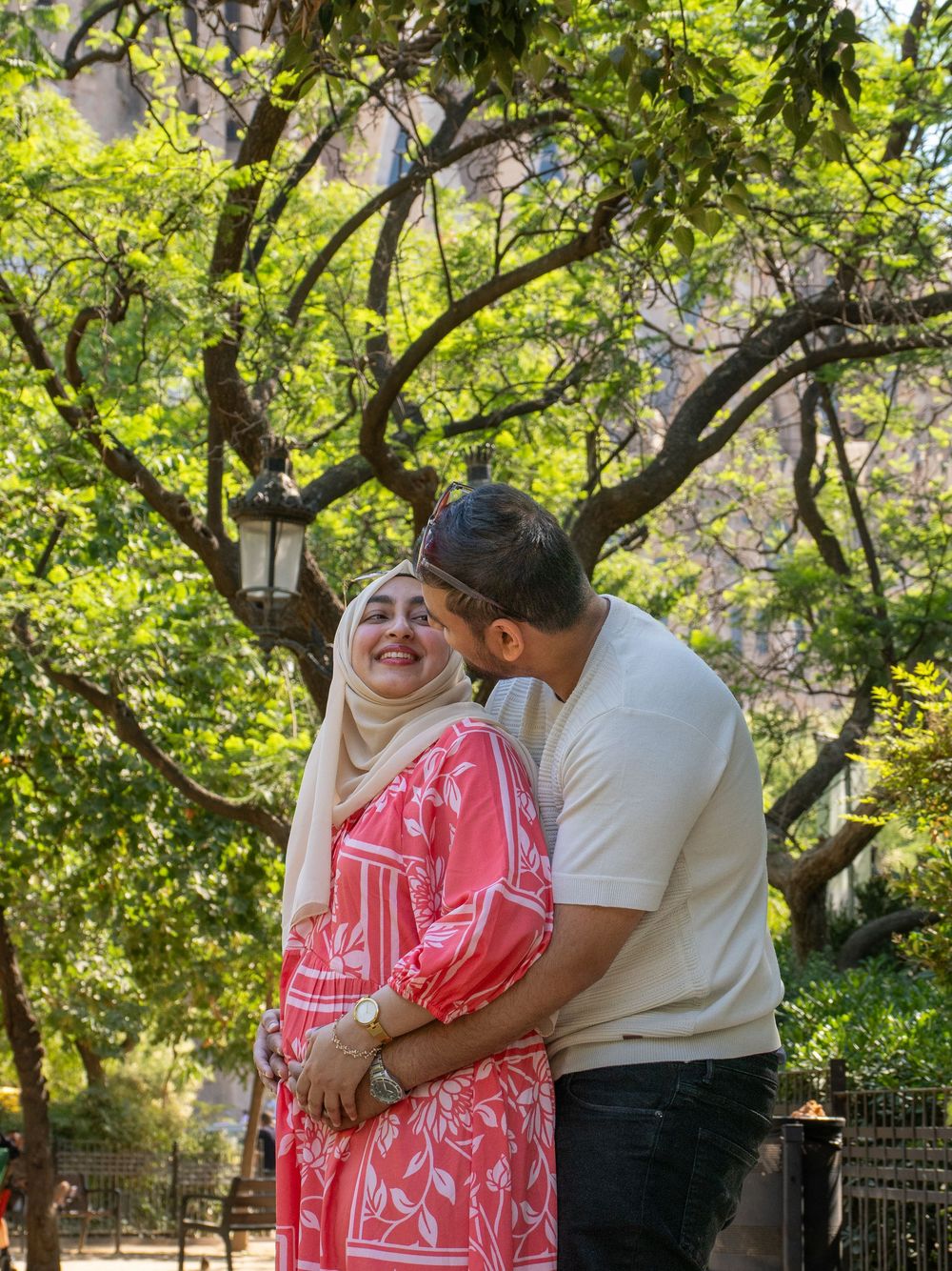 A couple sharing a tender moment outdoors, woman in pink floral dress and hijab, man in white tee, lush green trees behind.