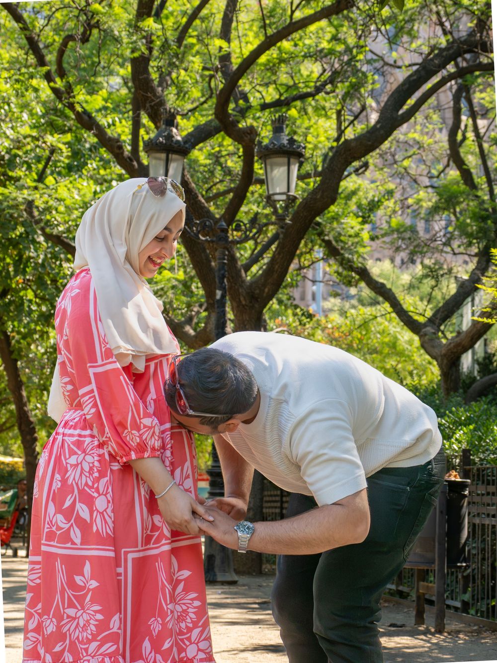 Man bowing to kiss woman's hand in pink floral hijab dress outdoors in lush green park setting.