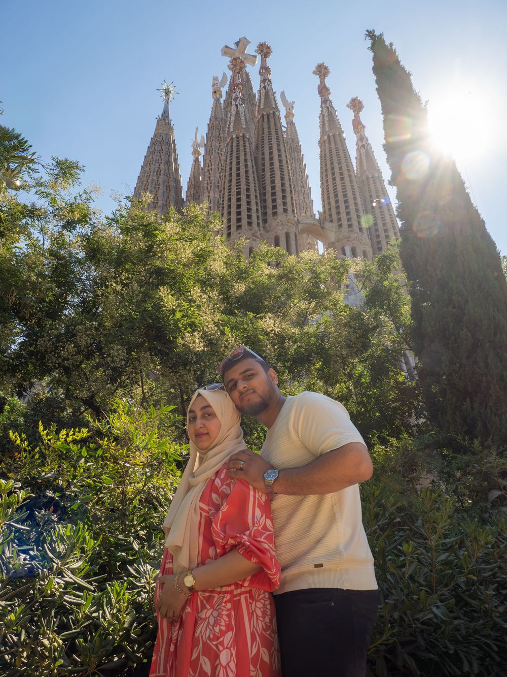 Couple posing in front of Sagrada Familia basilica in Barcelona, surrounded by lush green trees on a sunny day.