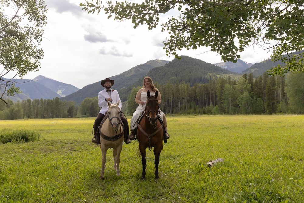 Two riders on horseback in a lush green mountain meadow surrounded by trees and scenic peaks.