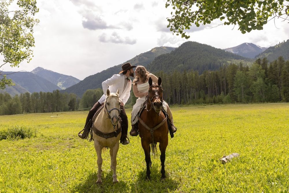 Two riders on horseback in a green meadow with mountain forest backdrop on a cloudy day.