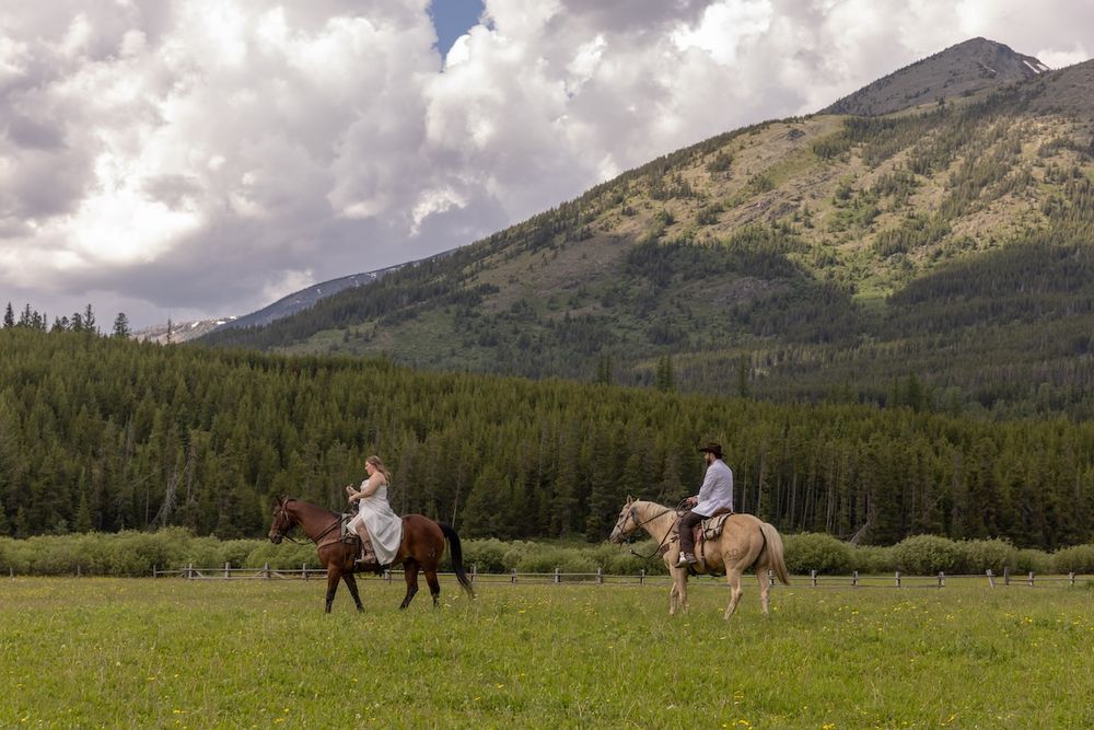 Two riders on horseback traverse a lush green meadow with a forested mountain backdrop under dramatic cloudy skies.