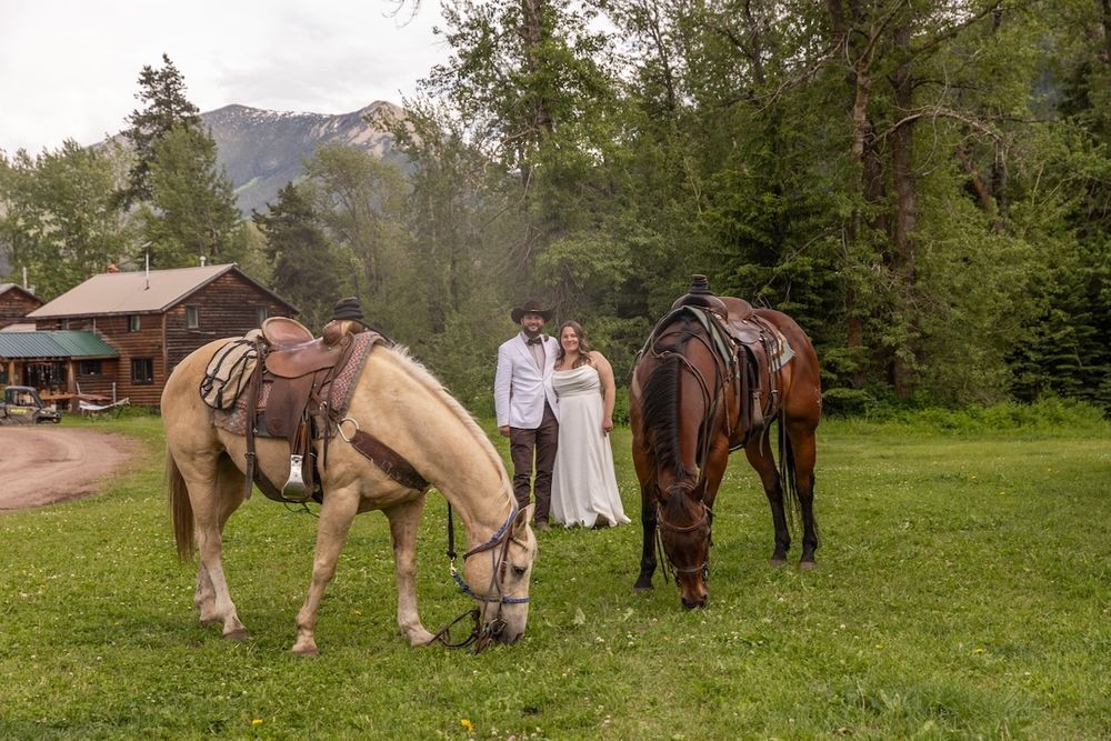 Bride and groom posing with two saddled horses on green meadow with rustic cabin and mountains backdrop.