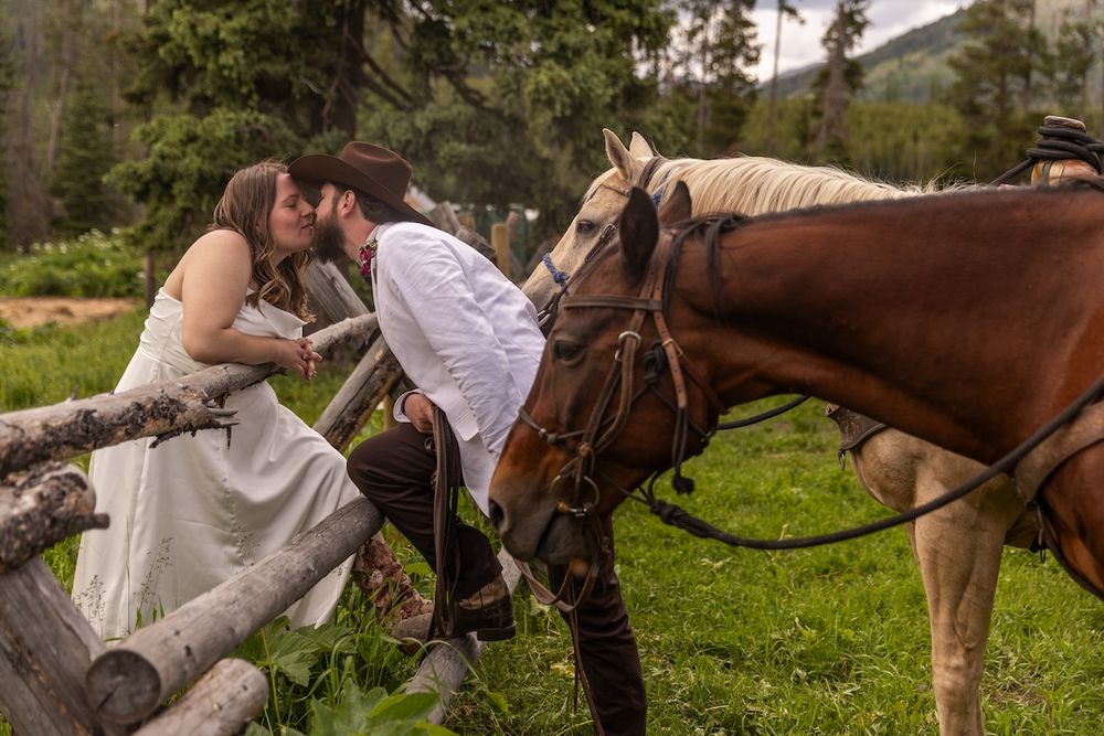Couple sharing a romantic kiss by a rustic fence with two horses in a lush green mountain meadow.