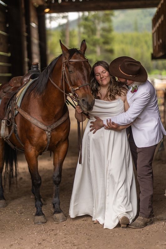 A bride and groom in western attire embrace tenderly beside a saddled brown horse inside a rustic barn.