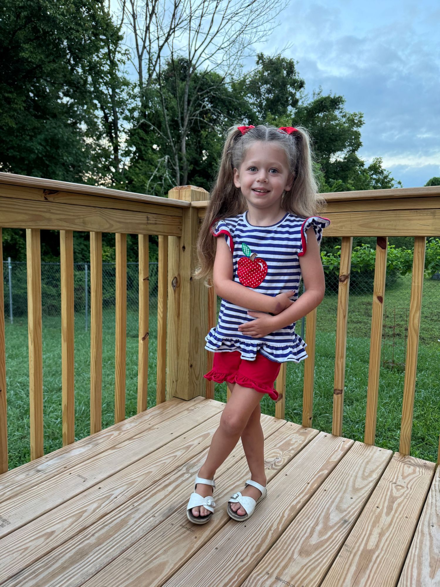 Young girl in striped top with apple applique and red shorts stands on wooden deck outdoors.
