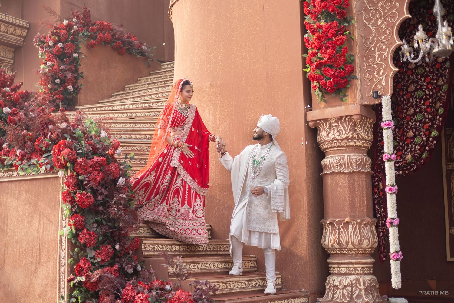 Indian bride in red lehenga and groom in white sherwani hold hands on ornate stairs adorned with red flowers.