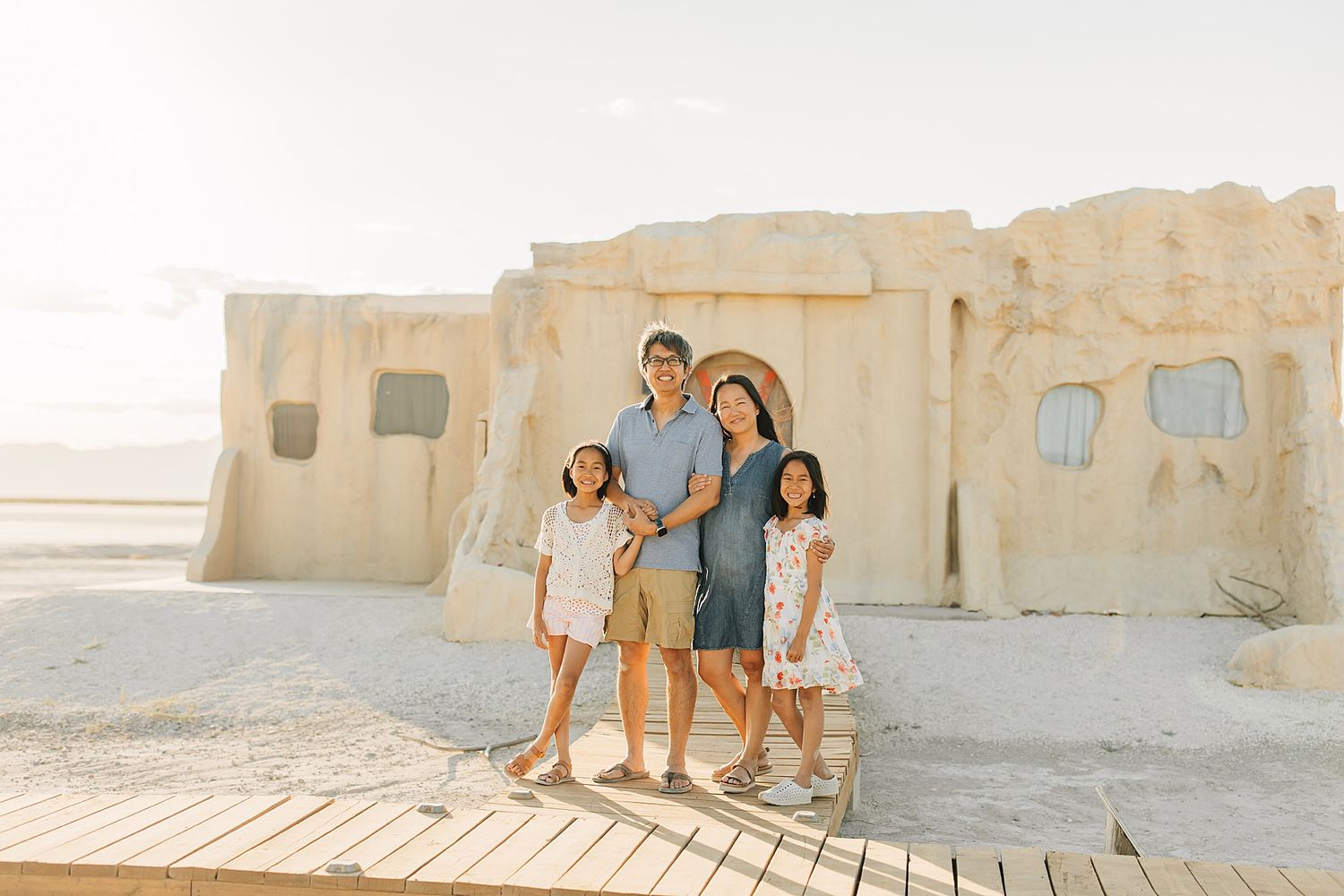 Family of four posing together outdoors near sandy adobe-style building in bright sunlight.