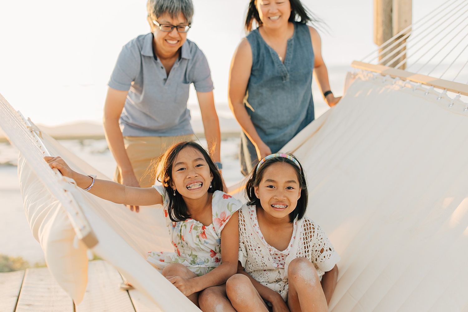 Two smiling girls sit in a hammock while two women stand behind them on a sunny outdoor deck.