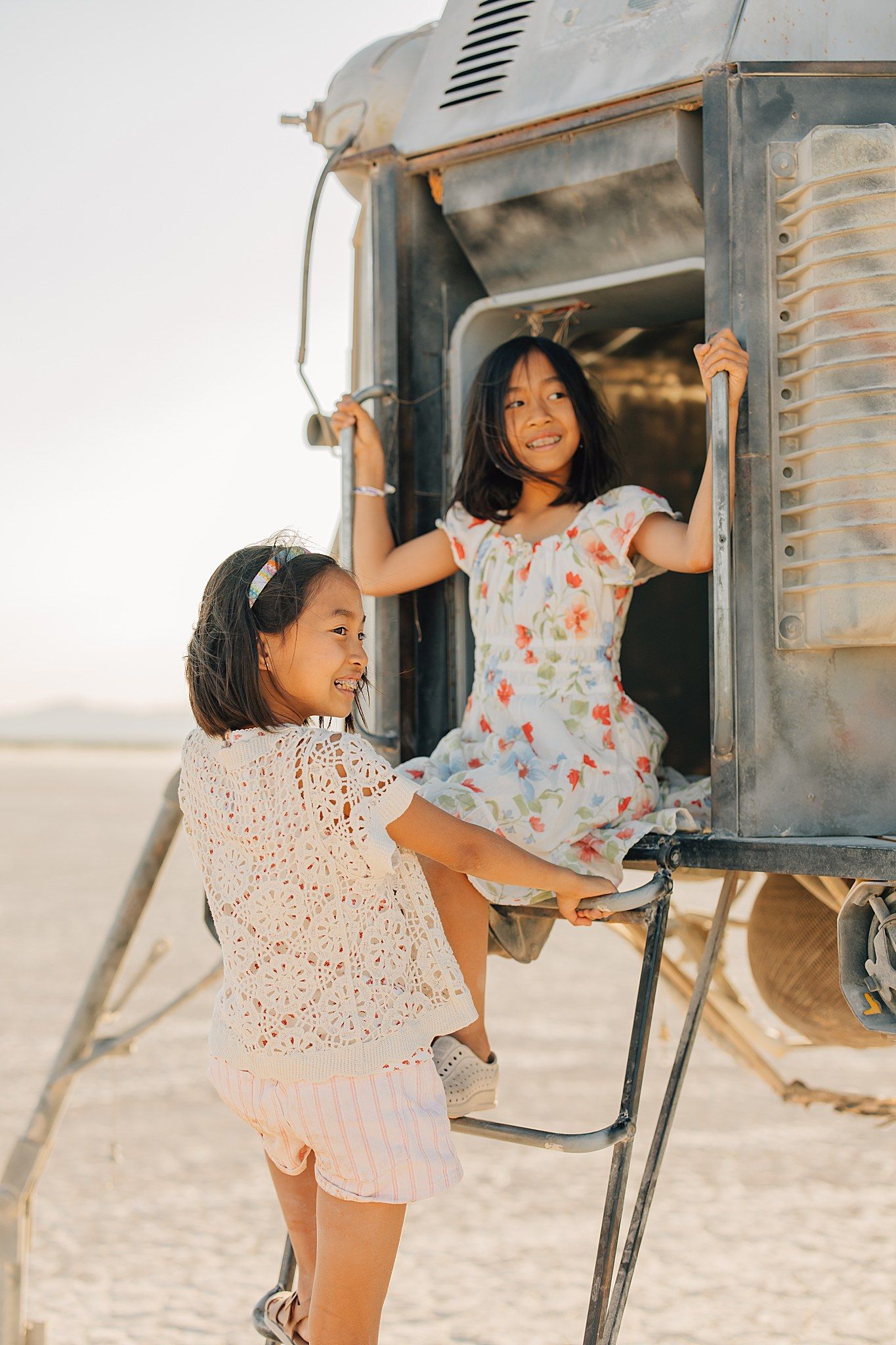 Two young girls in floral dresses playing on an old vintage vehicle in a sandy desert landscape.