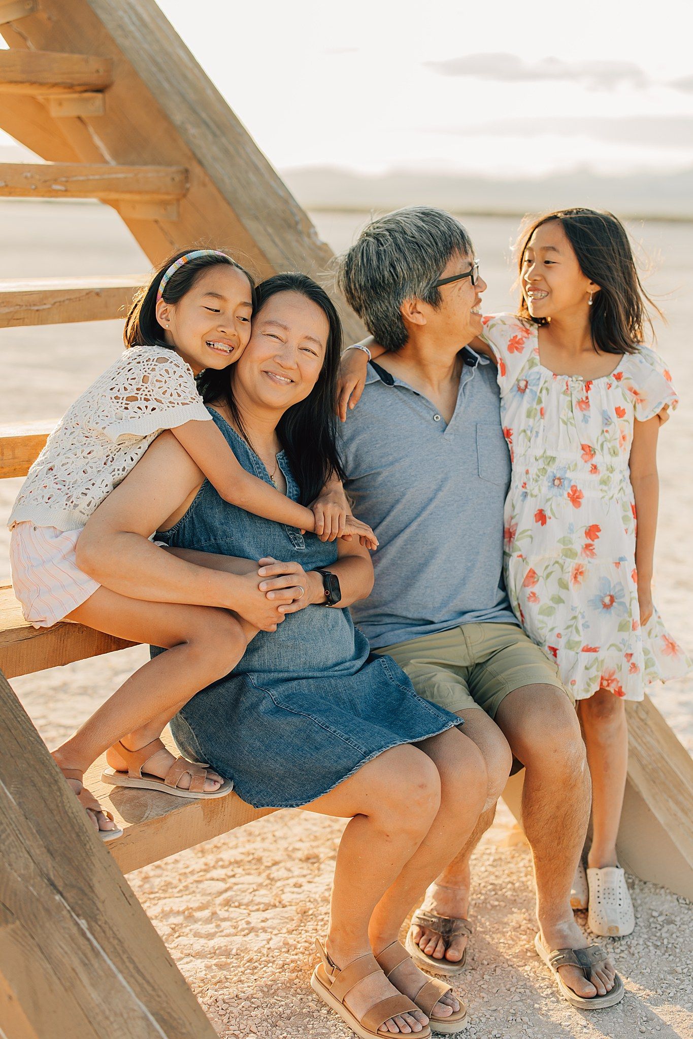 Happy Asian family of four smiling and hugging on a sunny beach, two daughters with parents sitting on wooden structure.
