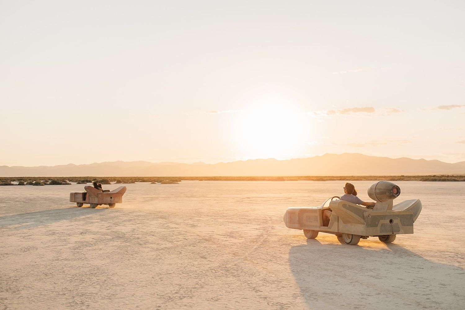 Two people sitting in lounge chairs on a vast salt flat, watching a golden sunset over distant mountains.