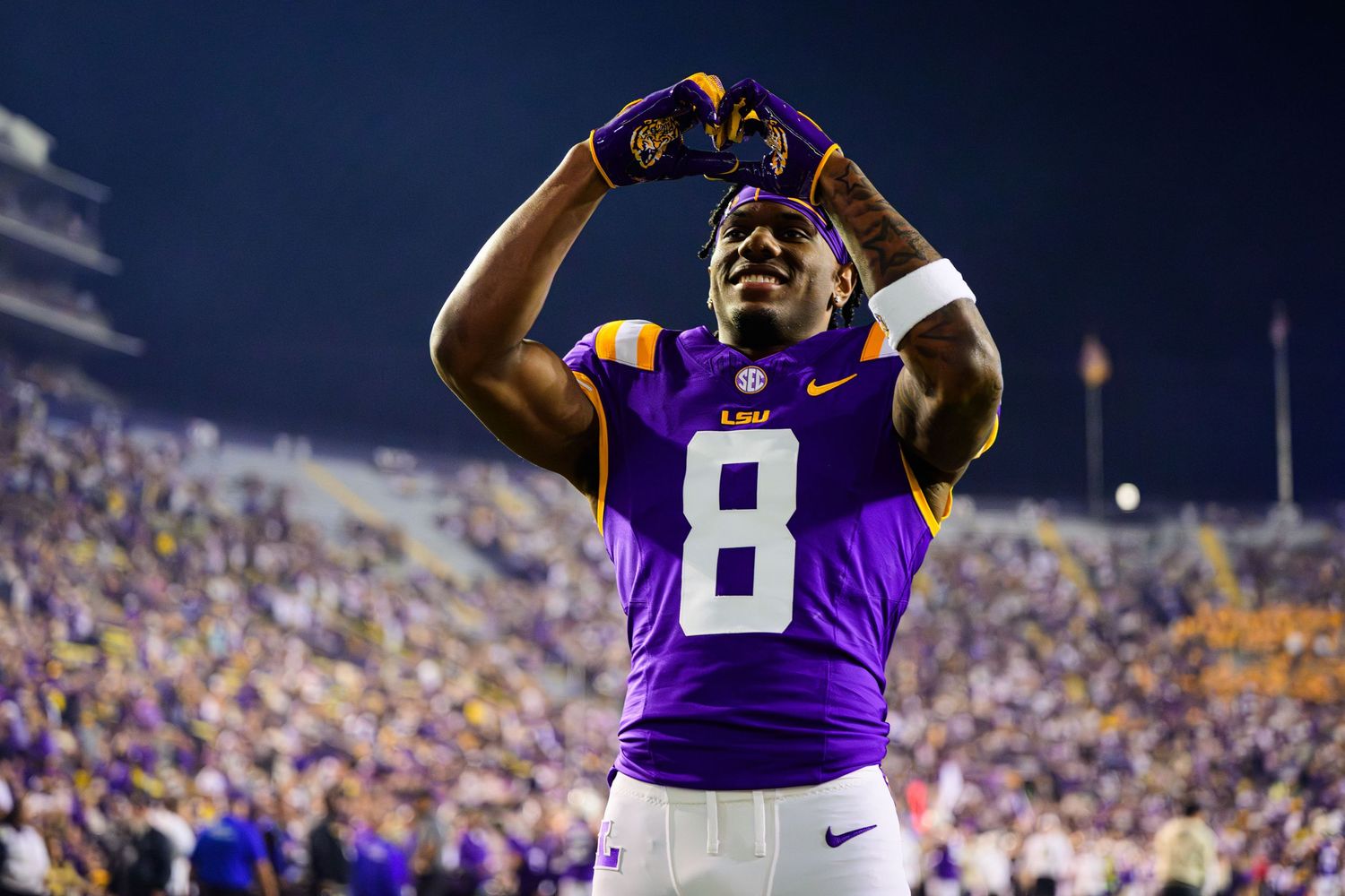 LSU Tigers football player wearing purple jersey number 8 celebrates on field at night game in packed stadium.