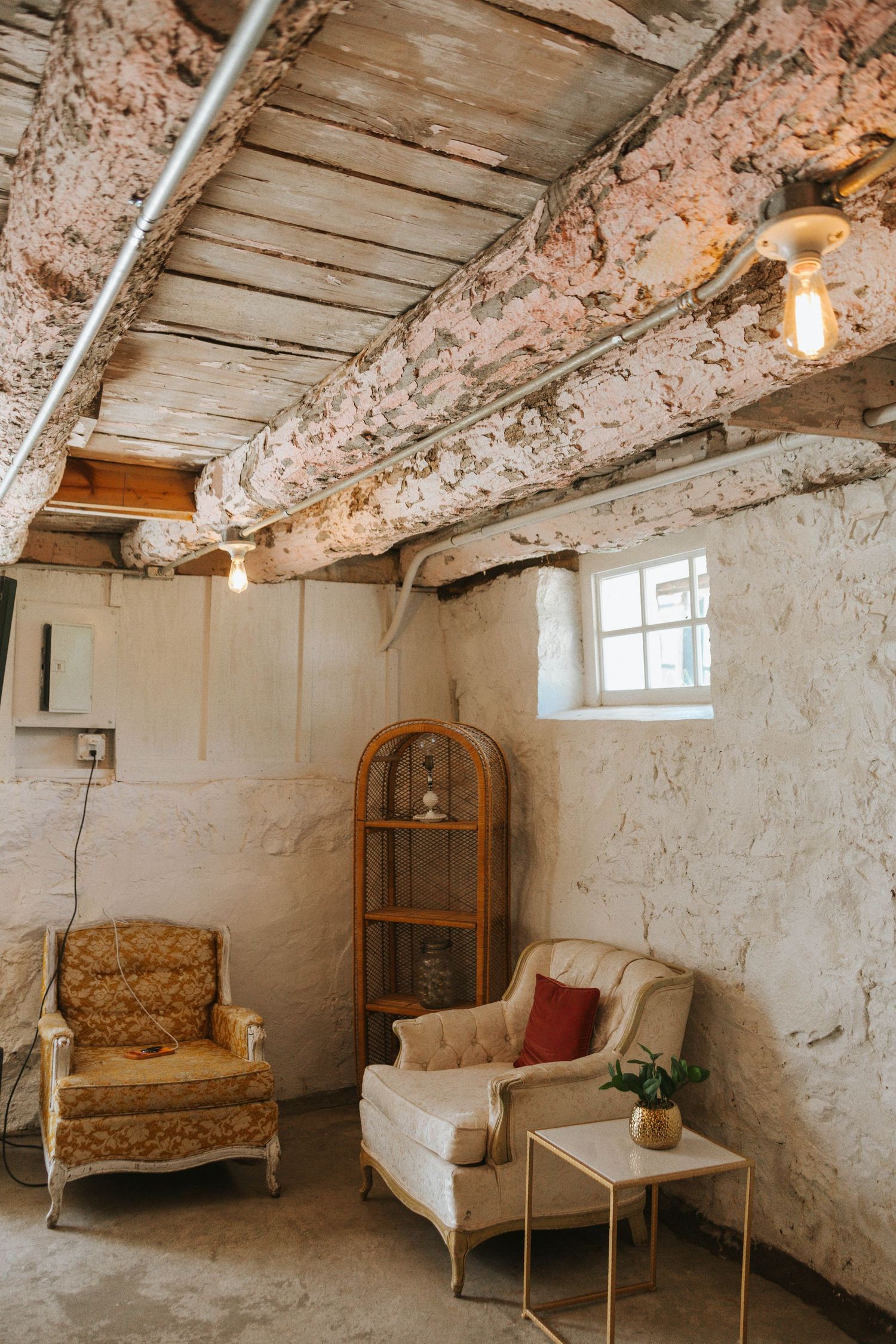 Rustic vintage sitting room with exposed wooden beams, peeling white walls, two antique chairs, and warm Edison bulb lighting.