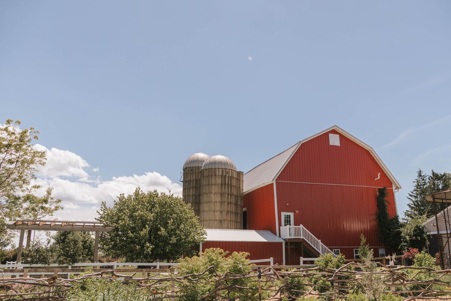 Red barn with silver silos surrounded by lush green trees under a bright blue sky on a sunny farm day.